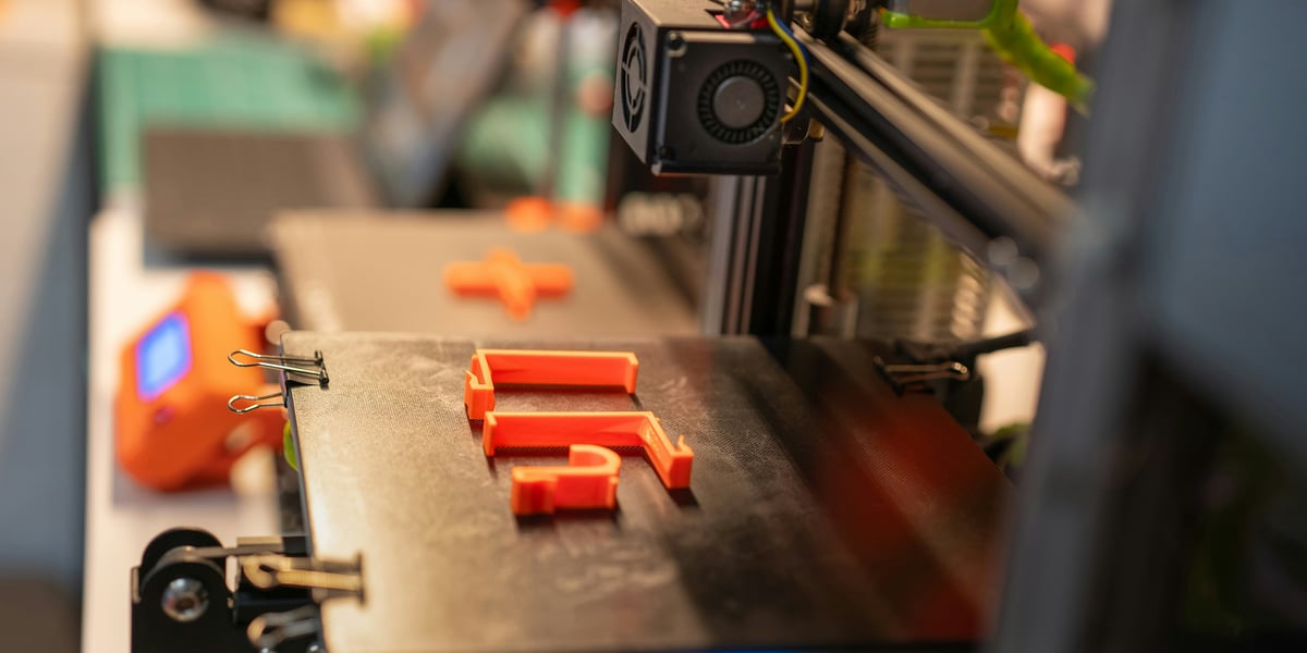 Na fotografii s mělkou hloubkou ostrosti je černá tisková podložka 3D tiskárny s částečně vytištěným oranžovým tvarem písmene „F" a oranžovým tvarem plus, zajištěnými kovovými sponami. V nezaostřeném pozadí jsou viditelné další prvky tiskárny, včetně ventilátoru a elektronických součástek. Okolní osvětlení naznačuje vnitřní prostředí, jako je dílna nebo kancelář.