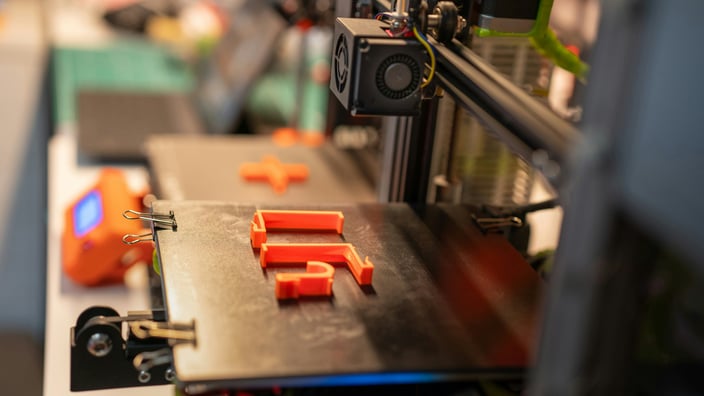 Na fotografii s mělkou hloubkou ostrosti je černá tisková podložka 3D tiskárny s částečně vytištěným oranžovým tvarem písmene „F“ a oranžovým tvarem plus, zajištěnými kovovými sponami. V nezaostřeném pozadí jsou viditelné další prvky tiskárny, včetně ventilátoru a elektronických součástek. Okolní osvětlení naznačuje vnitřní prostředí, jako je dílna nebo kancelář.