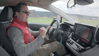 Человек в красном жилете за рулем Toyota Corolla, сфокусированный на дороге, интерьер автомобиля виден с приборной панелью и экраном мультимедиа.