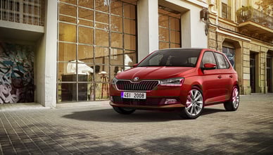 Красный хэтчбек Skoda Fabia третьего поколения с черной крышей припаркован перед современным зданием с большими стеклянными окнами, отражающими дневной свет. Здание имеет индустриальный вид с металлическими оконными рамами, а в интерьере видны растения и белая мебель. Тротуар вымощен плиткой, и слева виден фрагмент граффити. На заднем плане справа находится традиционное городское здание с балконами.