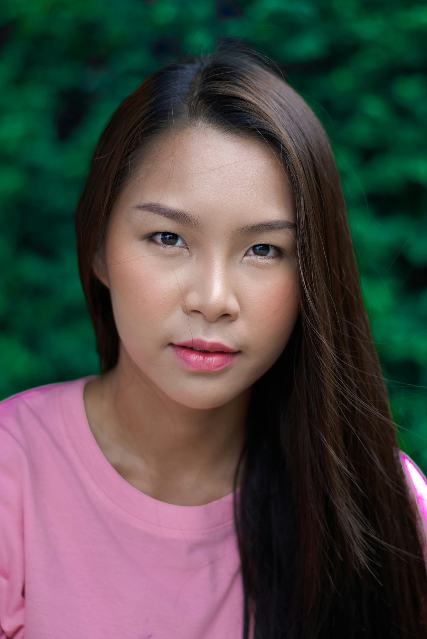 Portrait with natural makeup, pink top, long straight hair, green blurred background.
