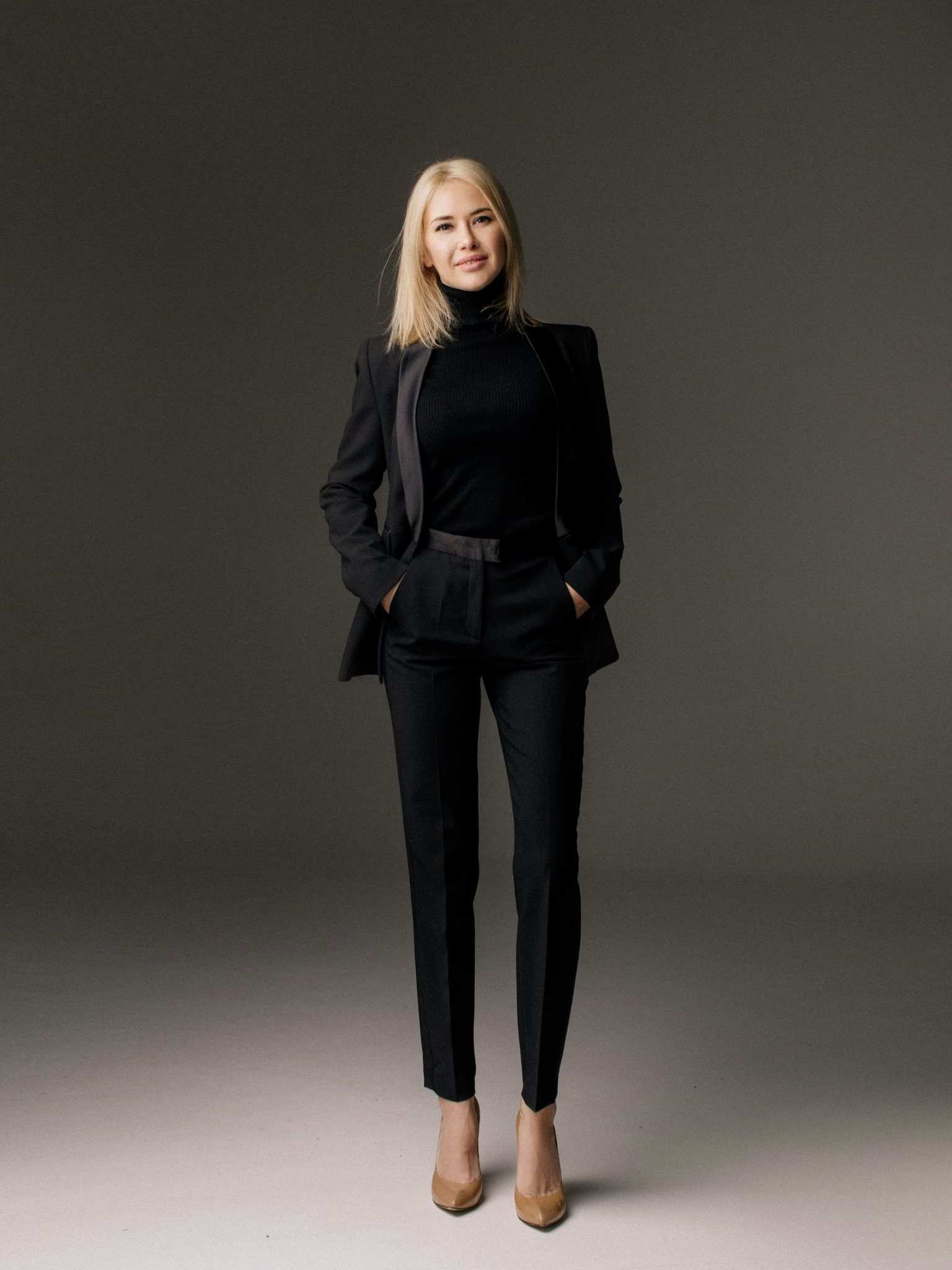 A woman in a black tailored suit with a turtleneck, standing confidently in a minimalist studio setting.