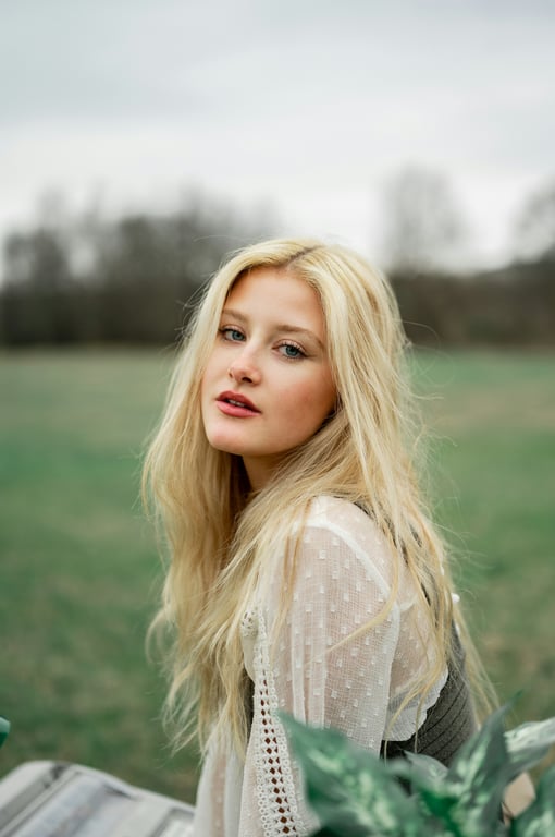 A woman outdoors with long blonde hair, wearing a light sheer blouse, posing against an open green field.