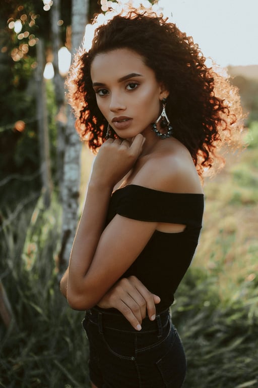 Woman in off shoulder black top posing outdoors at sunset with curly hair and soft golden light behind her.