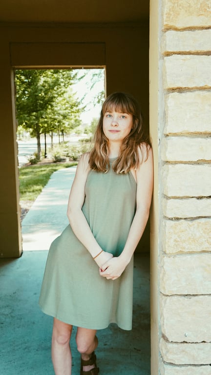 The subject stands outdoors in a loose green dress near a stone wall.