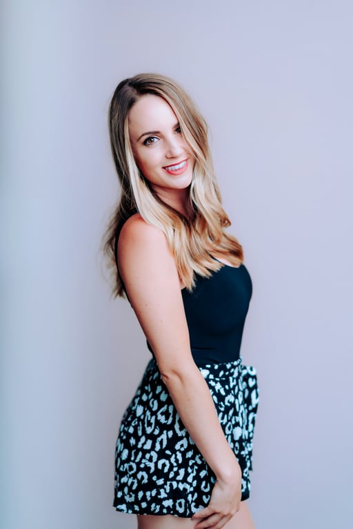 Smiling woman in a sleeveless black top and patterned shorts posing against a soft light studio background.