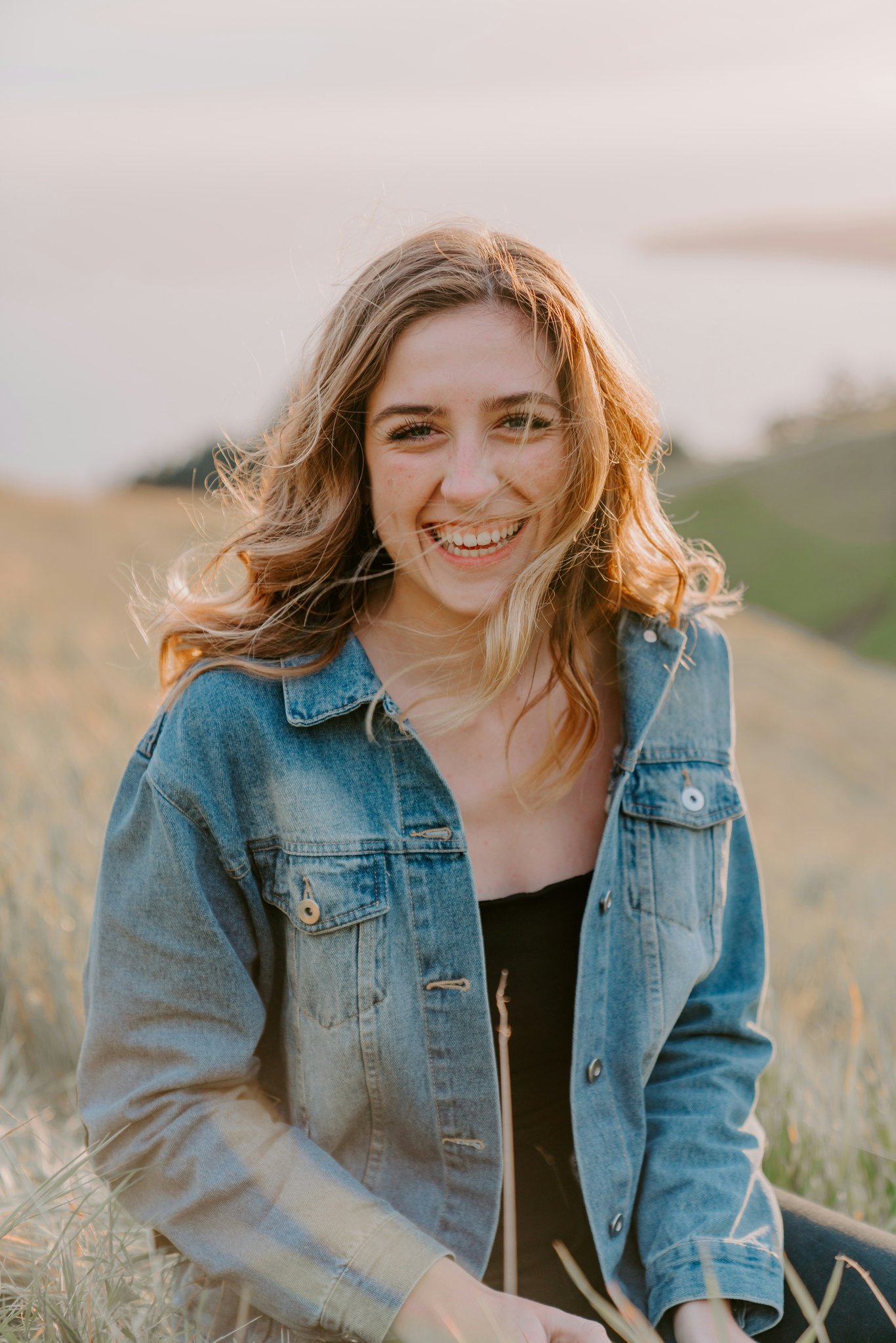 A woman outdoors wearing a denim jacket, smiling in natural light with grassy hills and soft sunset tones behind her.