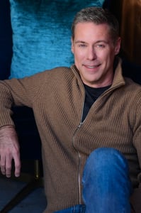 leaning against a teal fabric, Roy smiles at the camera, wearing a light brown zipper sweater over a dark T.