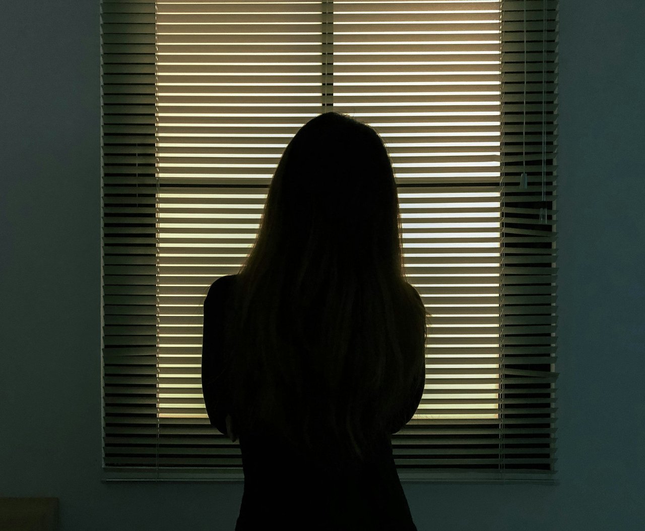 Silhouette of a young woman standing in front of a window with the blinds half open