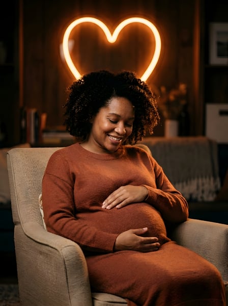 MULHER GRÁVIDA SEGURANDO A BARRIGA SENTADA NO SOFÁ COM UM CORAÇÃO LUMINOSO AO FUNDO 1