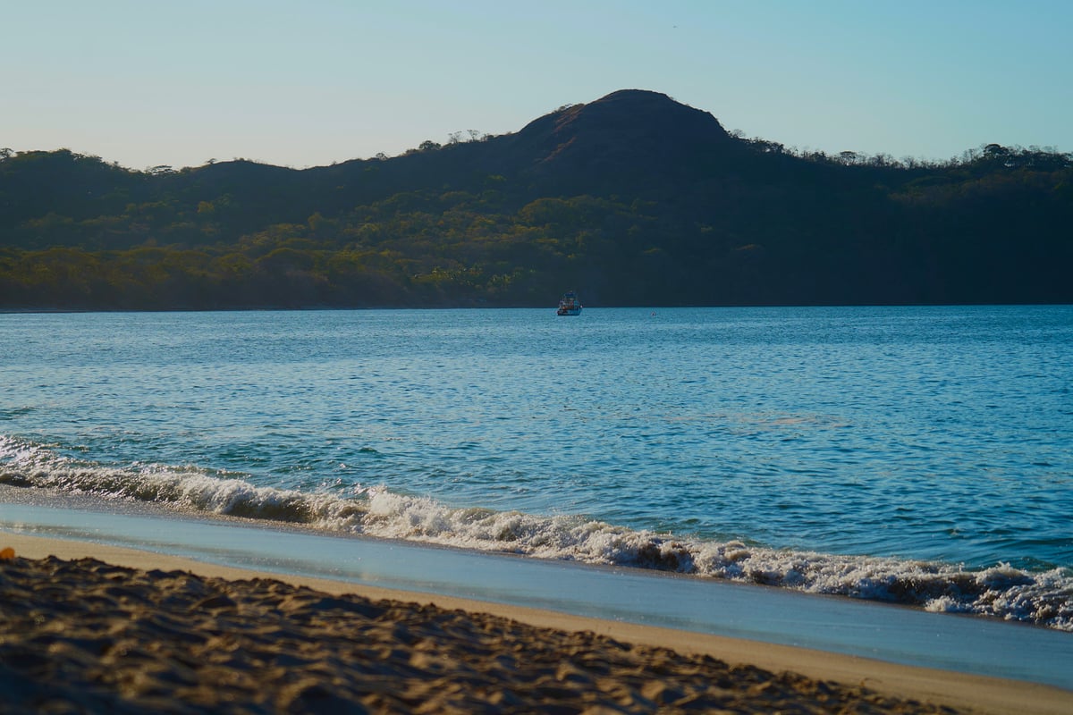 A small boat floats in a calm blue bay beneath a large mountain as gentle waves break on a sandy beach under soft afternoon light.