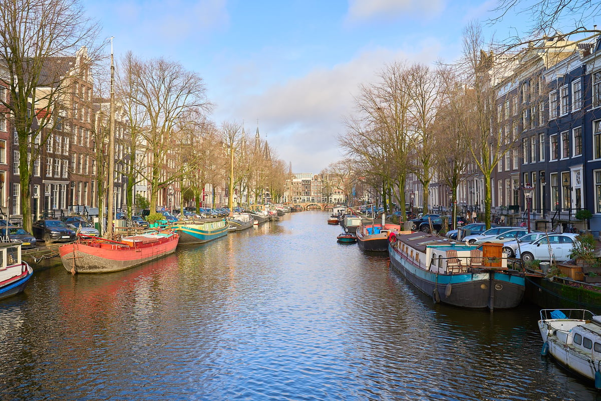 A wide canal in Amsterdam is lined with colorful houseboats and historic brick buildings under a bright blue sky with bare winter trees.