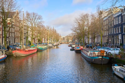 A wide canal in Amsterdam is lined with colorful houseboats and historic brick buildings under a bright blue sky with bare winter trees.