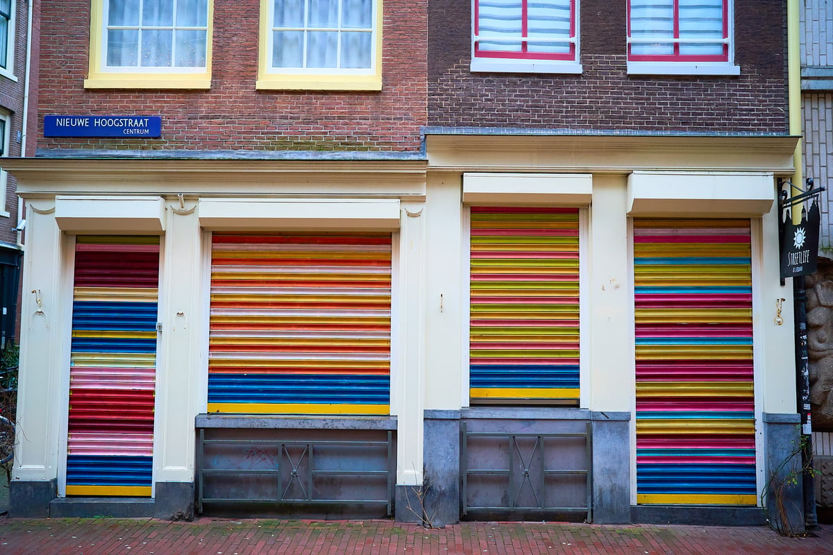 Four storefronts with brightly colored horizontal striped metal shutters are set into the ground floor of a brick building on Nieuwe Hoogstraat in Amsterdam.