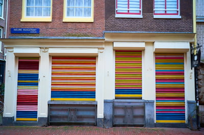 Four storefronts with brightly colored horizontal striped metal shutters are set into the ground floor of a brick building on Nieuwe Hoogstraat in Amsterdam.