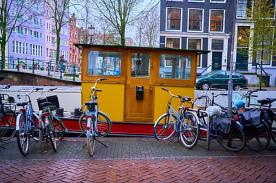 A yellow wooden canal boat cabin sits behind a row of parked bicycles on a brick sidewalk in Amsterdam under an overcast sky.