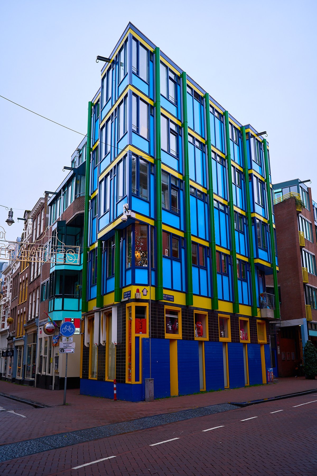 A tall, modern building painted in bright blue, yellow, and green stands on a cobblestone street corner in Amsterdam under an overcast sky.