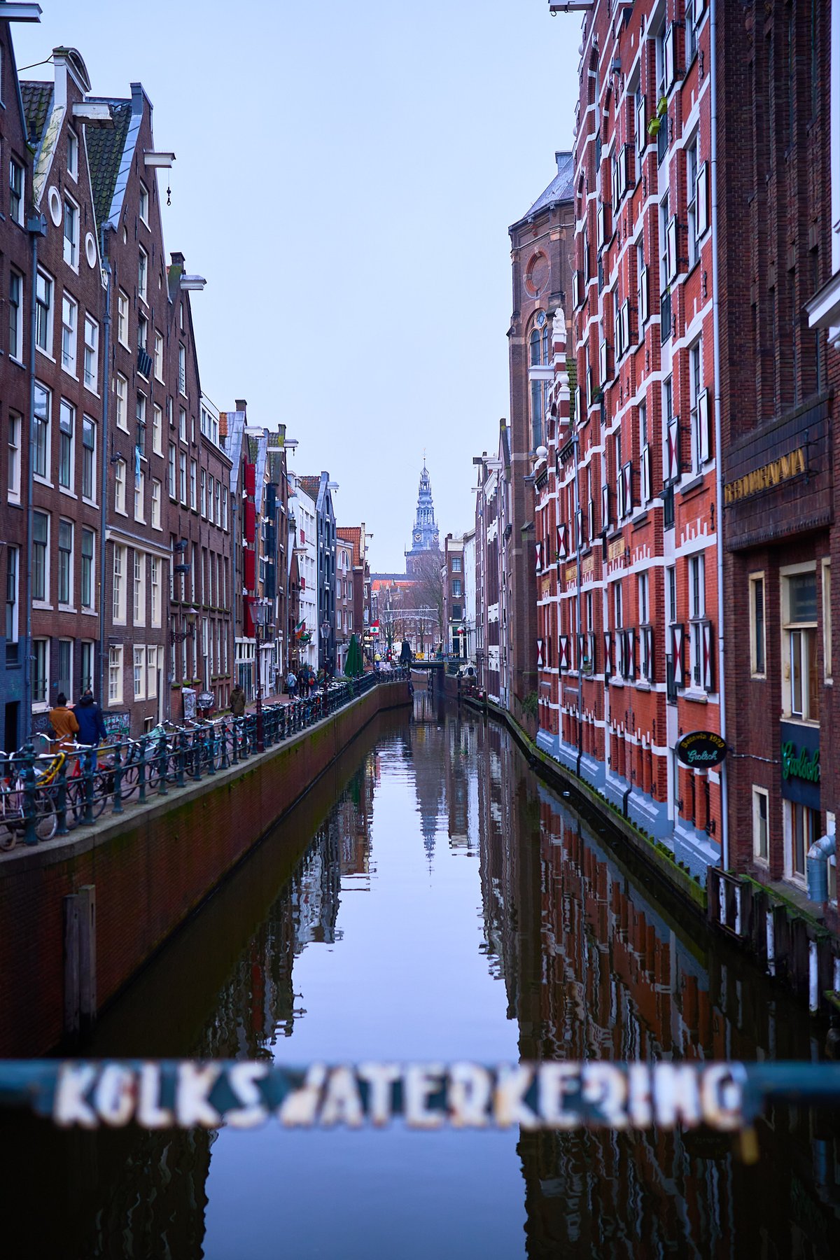 A narrow canal in Amsterdam is lined with tall brick buildings and leads toward a distant church spire under a soft blue sky.
