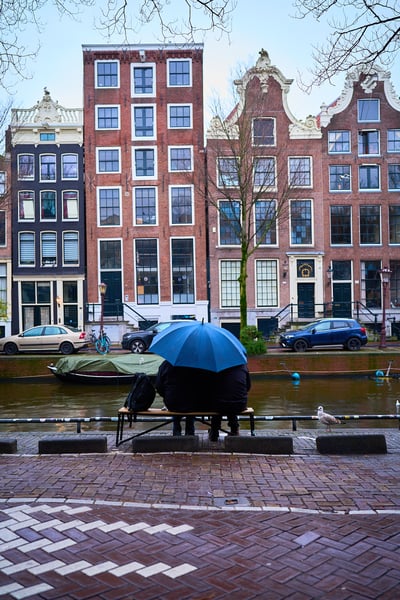 An elderly couple sit on a bench under a bright blue umbrella facing a canal lined with traditional Dutch architecture on an overcast day.