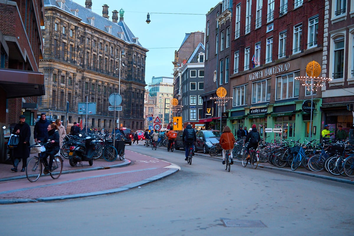 Cyclists and pedestrians navigate a busy street in Amsterdam at dusk, flanked by historic brick buildings and rows of parked bicycles.