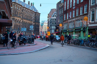 Cyclists and pedestrians navigate a busy street in Amsterdam at dusk, flanked by historic brick buildings and rows of parked bicycles.