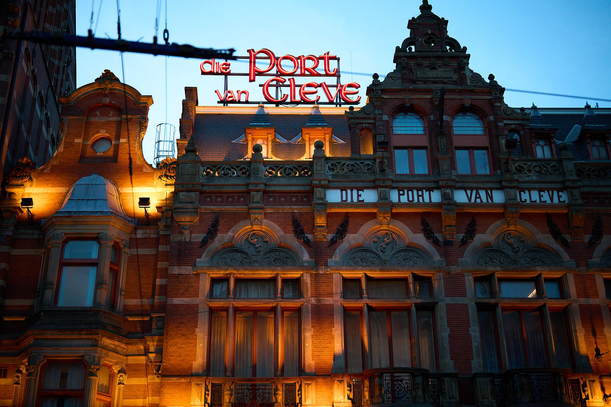 The ornate brick facade of the Die Port van Cleve hotel in Amsterdam is illuminated by warm spotlights and a red neon sign against a twilight sky.