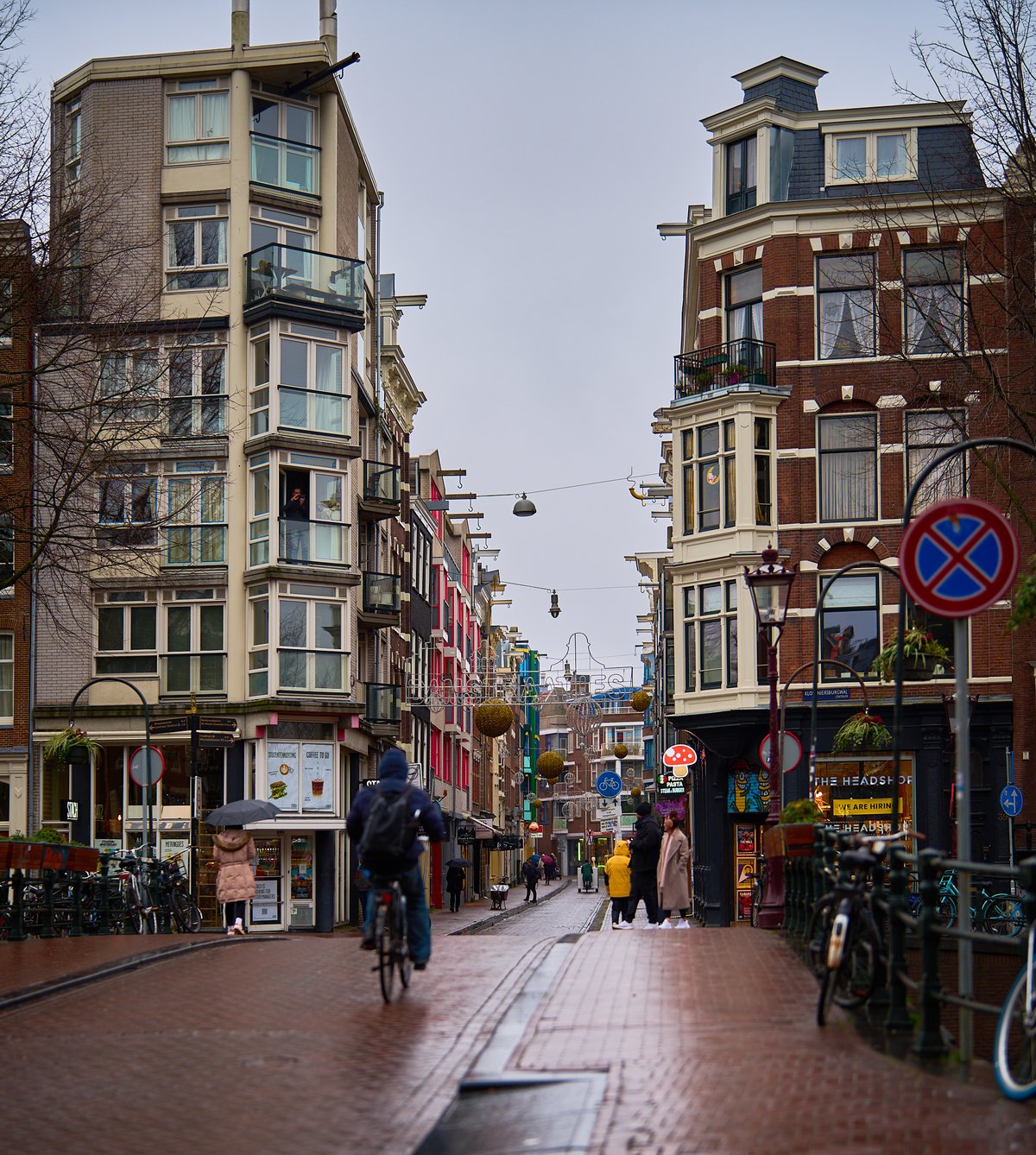 A cyclist pedaling away down a wet brick street lined with tall, narrow buildings under a grey, overcast sky in Amsterdam.
