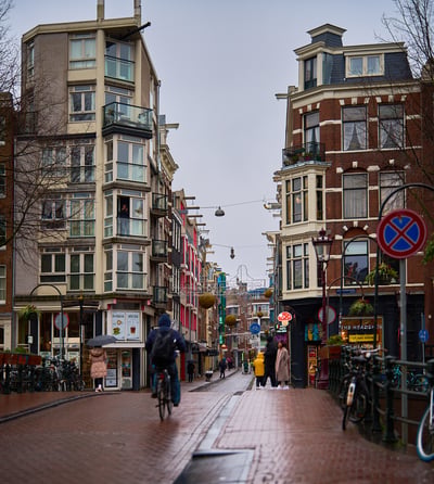 A cyclist pedaling away down a wet brick street lined with tall, narrow buildings under a grey, overcast sky in Amsterdam.