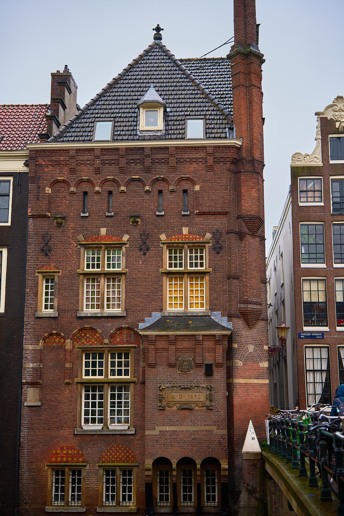 A historic red brick building with ornate masonry and a date plaque from 1890 stands beside a canal bridge lined with bicycles in Amsterdam.