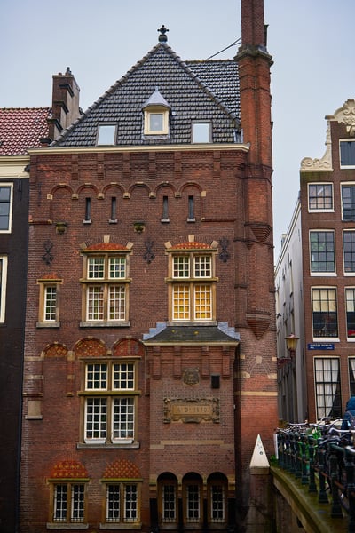 A historic red brick building with ornate masonry and a date plaque from 1890 stands beside a canal bridge lined with bicycles in Amsterdam.