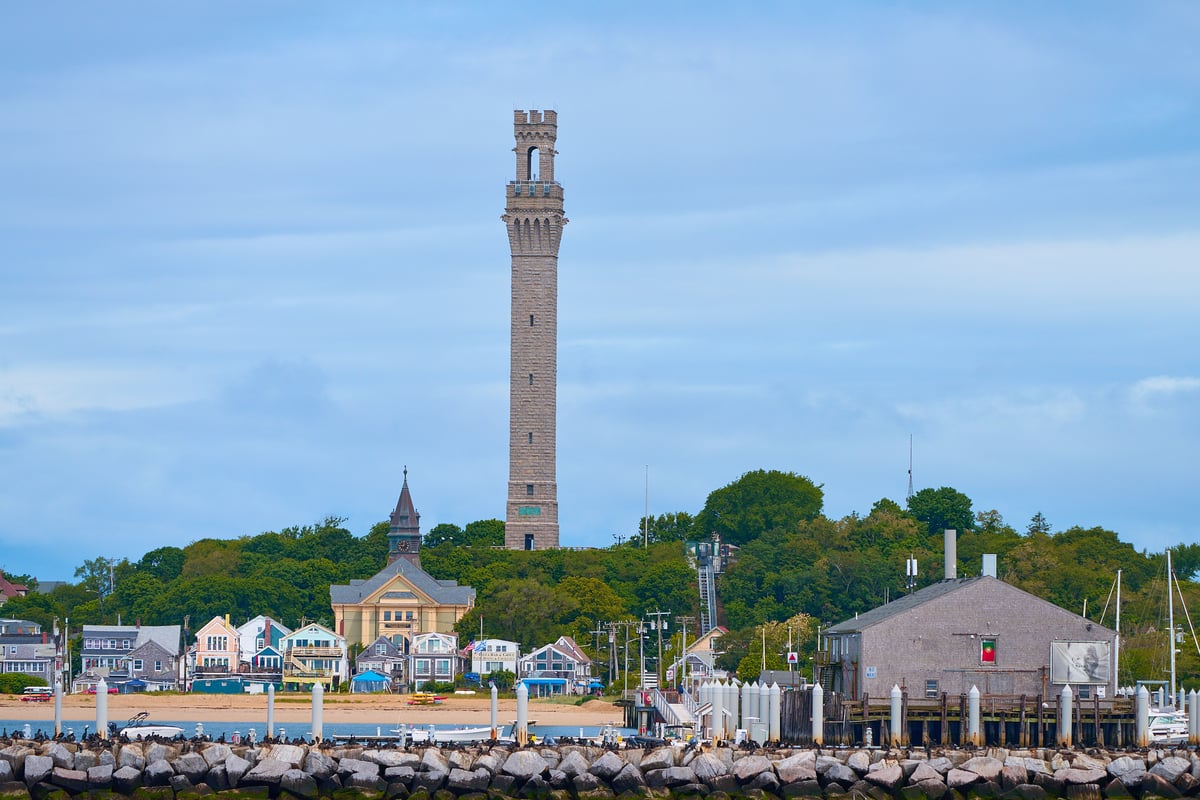 A tall granite monument towers over a coastal town with colorful houses, a church, and a rock breakwater under an overcast sky.