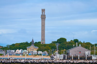 A tall granite monument towers over a coastal town with colorful houses, a church, and a rock breakwater under an overcast sky.