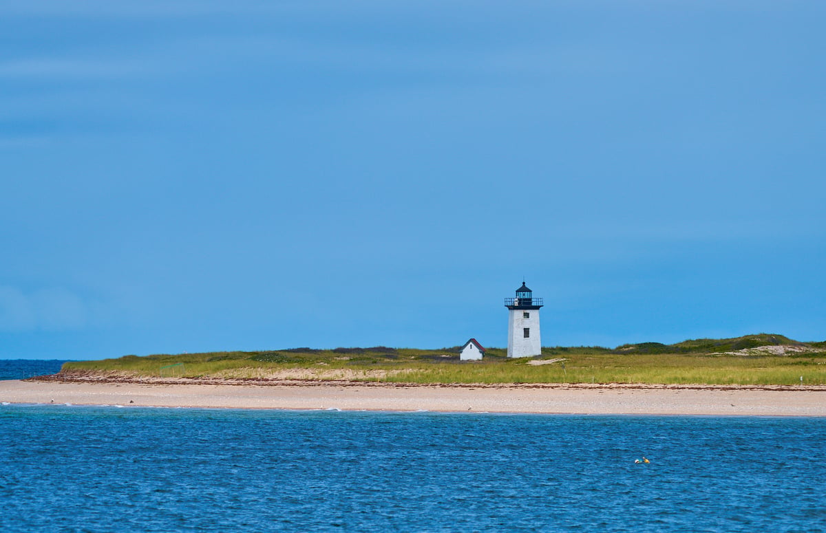 A white square lighthouse and a small outbuilding sit on a grassy sandbar between a calm blue sea and a clear sky.