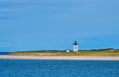 A white square lighthouse and a small outbuilding sit on a grassy sandbar between a calm blue sea and a clear sky.