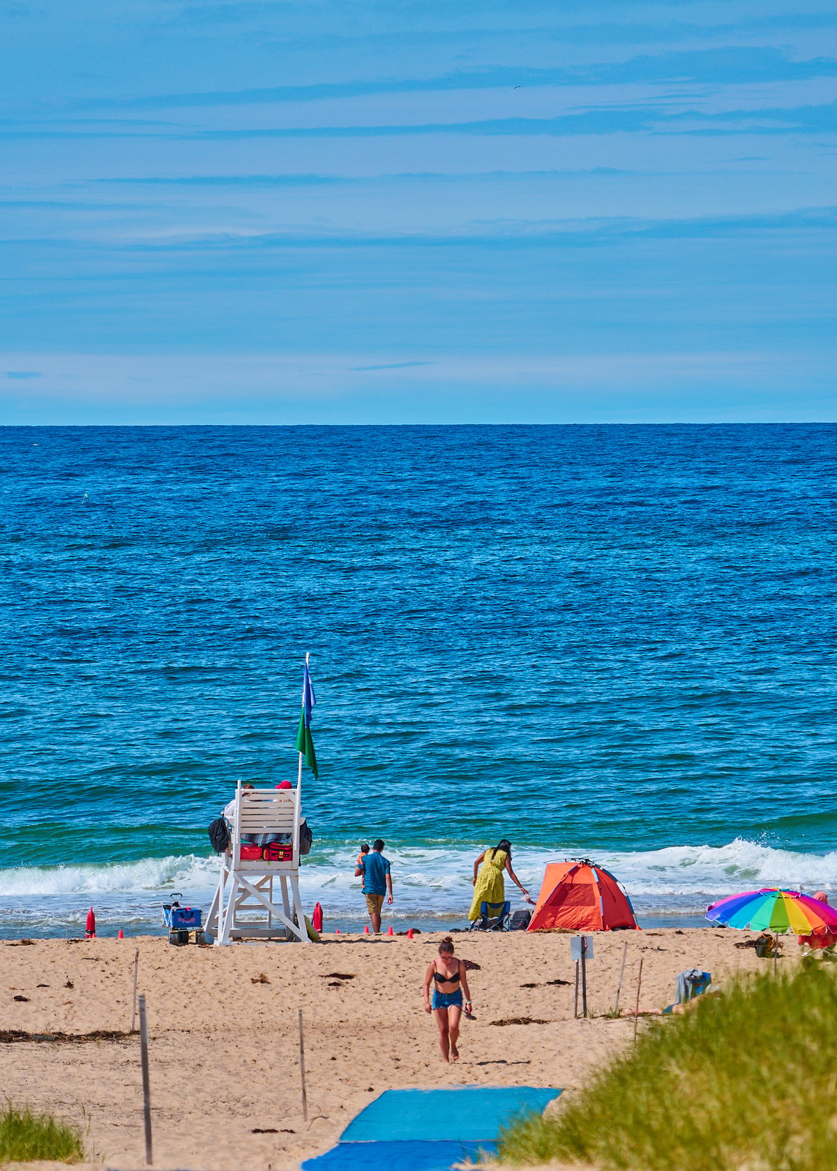 A white lifeguard stand stands on a sandy beach near people, an orange tent, and a multi-colored umbrella facing a blue ocean.