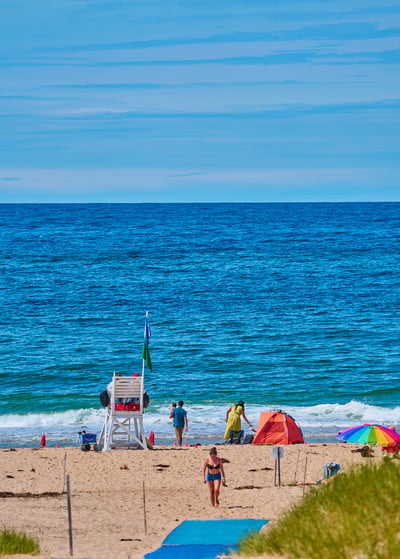 A white lifeguard stand stands on a sandy beach near people, an orange tent, and a multi-colored umbrella facing a blue ocean.