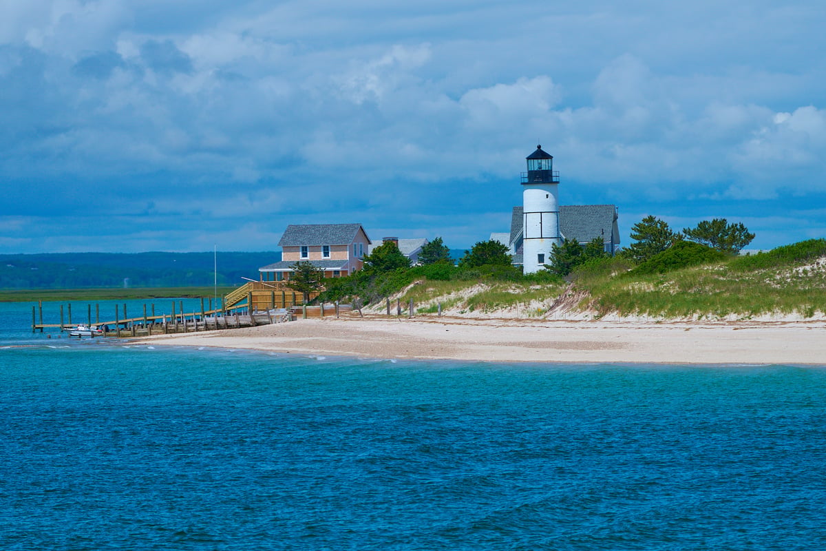 A white lighthouse and a shingled house stand on grassy dunes overlooking a sandy beach and blue water under a cloudy sky.