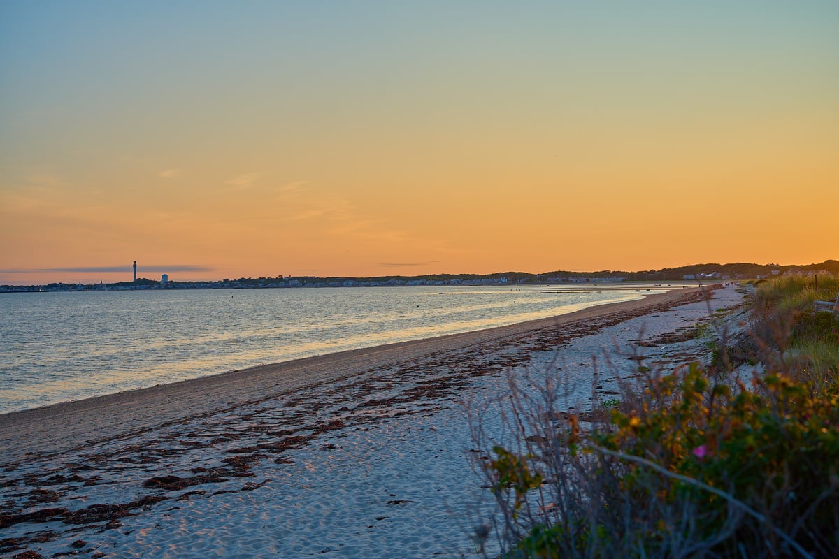 A sandy beach with lines of seaweed leads toward a distant town and tower under a glowing orange sunset sky.