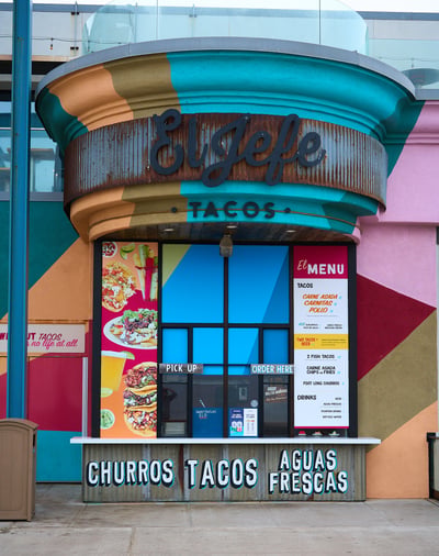 A vibrant taco stand named El Jefe features geometric color blocks, a corrugated metal sign, and a detailed menu board under bright daylight.