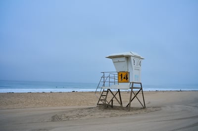 A white lifeguard tower numbered 14 stands on a wide sandy beach under a hazy blue sky with the ocean in the distance.