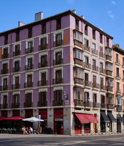 A multi-story purple building with ornate balconies and a street-level cafe sits on a sunny corner under a clear blue sky.