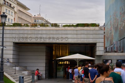 A crowd of people queues at the stone entrance of the Museo del Prado in Madrid beneath a large white umbrella.