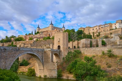 A medieval stone arch bridge spans a river leading to a fortified gatehouse and the Alcázar fortress on a hillside in Toledo, Spain.