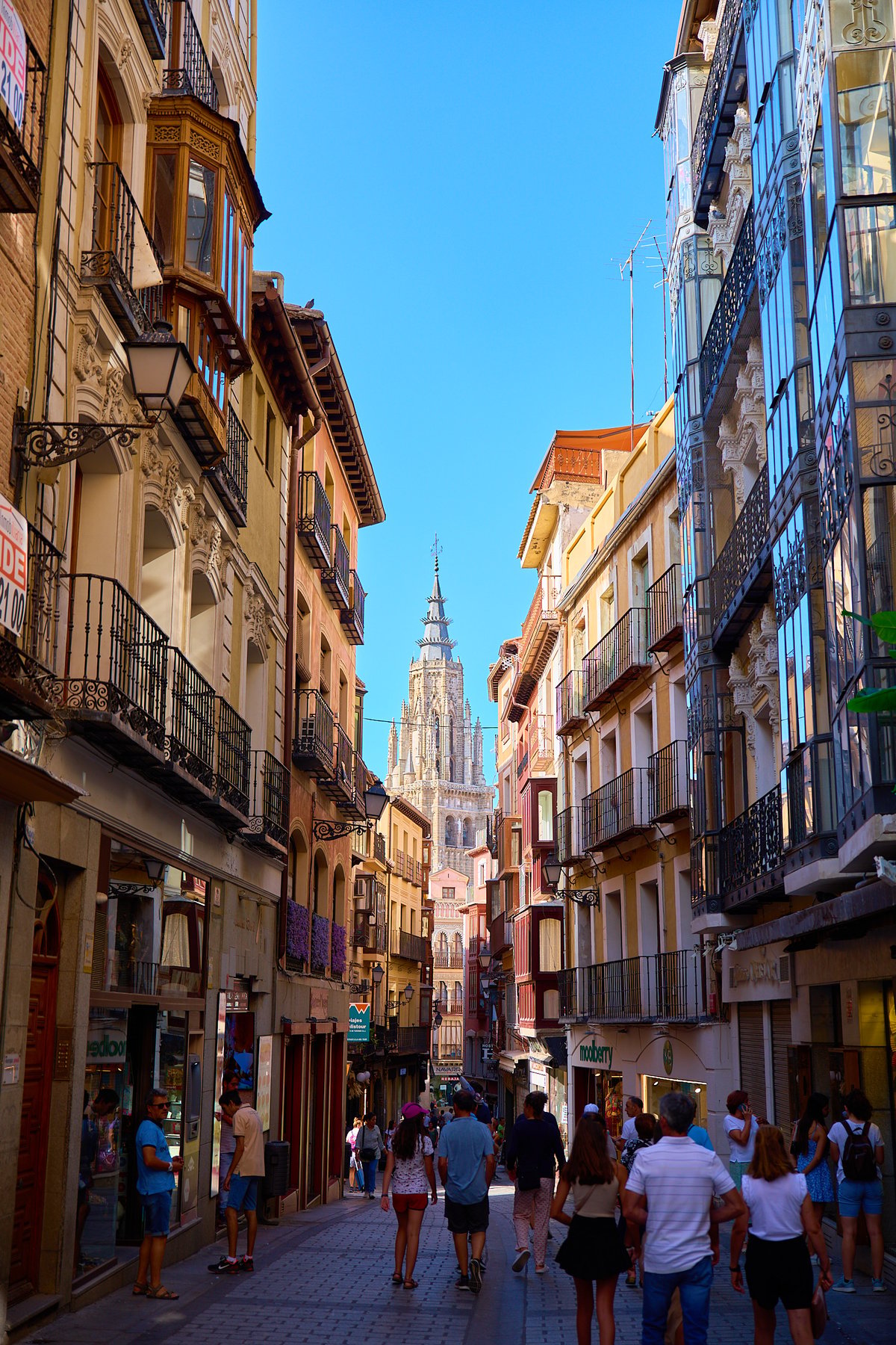A narrow cobblestone street lined with historic multi-story buildings leads toward a tall Gothic cathedral spire under a clear blue sky.