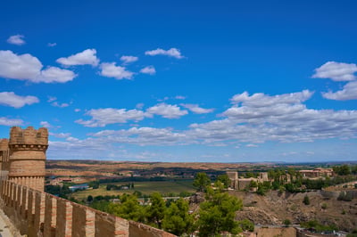 A view from a crenellated brick castle wall overlooking a green valley with a distant stone fortress under a bright blue sky with white clouds.