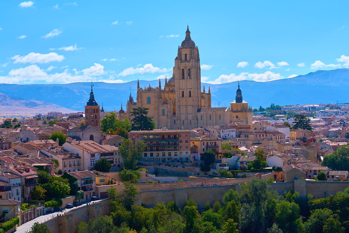 The Gothic Segovia Cathedral towers over a dense cityscape of terracotta-roofed buildings against a backdrop of blue mountains under a clear sky.