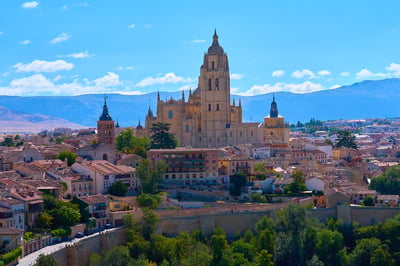 The Gothic Segovia Cathedral towers over a dense cityscape of terracotta-roofed buildings against a backdrop of blue mountains under a clear sky.