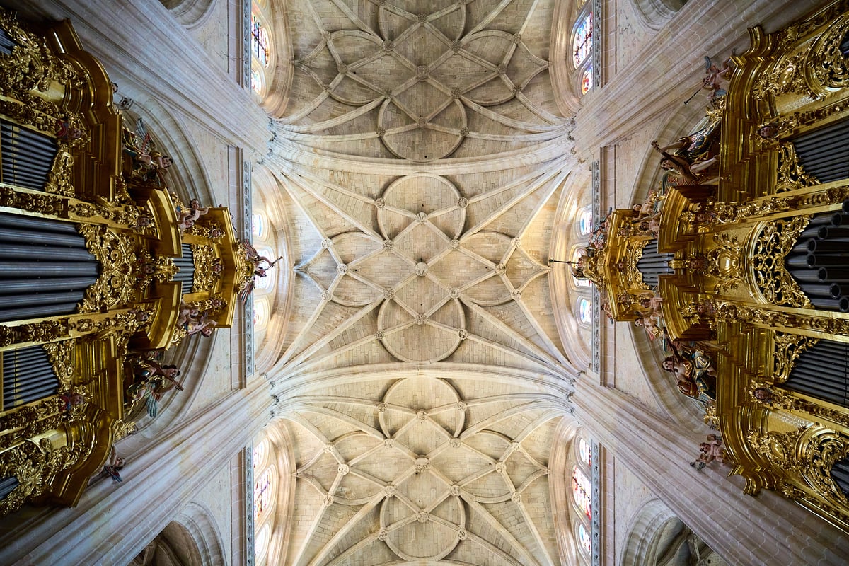 A low-angle perspective looks up at an intricate stone vaulted ceiling framed by two ornate golden pipe organs and stained glass windows.