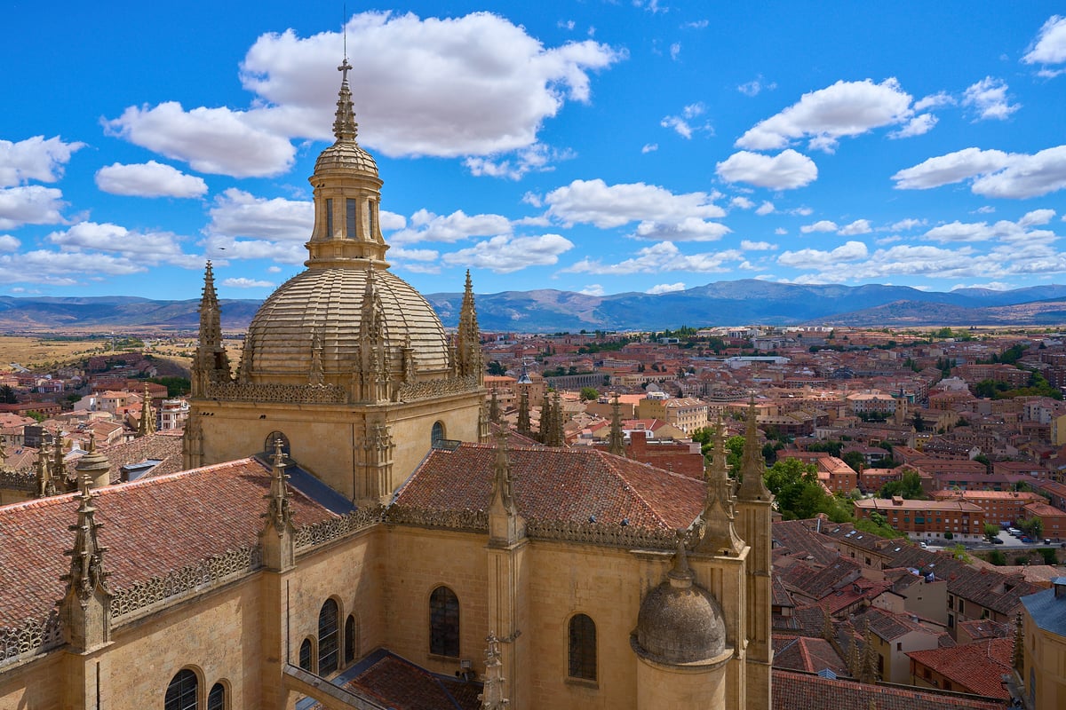 A high-angle view of the ornate stone dome and spires of Segovia Cathedral overlooking a city of terracotta roofs and distant mountains under a blue sky.