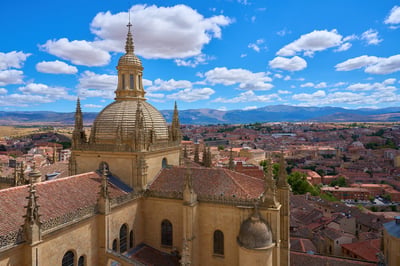 A high-angle view of the ornate stone dome and spires of Segovia Cathedral overlooking a city of terracotta roofs and distant mountains under a blue sky.